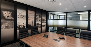 Modern boardroom featuring a large wooden meeting table, black leather chairs, NIZARRA designedAfrican wildlife acoustic wall panels, and desert-inspired sandblast film on glass partitions.