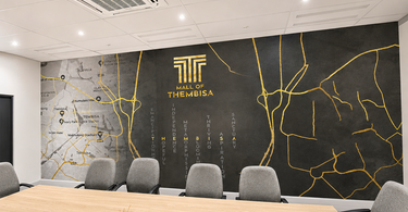 A modern boardroom with a custom Mall of Thembisa feature wall showing a grey map graphic, gold route lines, branded logo, and charcoal mural surface.