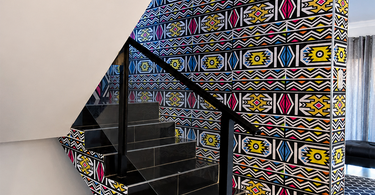 Wide view of a residential staircase transformed with bold Ndebele-inspired wallpaper across the stairwell walls and side panels.