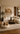 Wide-angle minimalist living and dining space featuring a cream L-shaped sectional sofa, wooden coffee table, dog on a cushion,neutral abstract wall art, and an indoor tree, with warm wood finishes and soft natural light creating a calm, refined atmosphere.