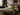 Olive-green NIZARRA Sepedi-inspired geometric cushion styled on a layered bed in an industrial bedroom with exposed brick, concrete walls and black metal details.
