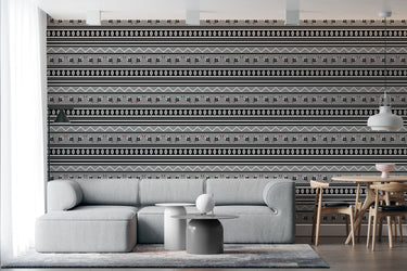 Swazi inspired wallpaper grey background with geometric repeat pattern of zig zag lines and shield and spear graphics and oval shapes in colours black and white. Setting is a living room with a corner sofa that complement the wallpaper.