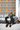NIZARRA Zulu-inspired geometric wallpaper with repeating multicoloured diamond and triangle patterns behind a grey leather sofa in a contemporary lounge with a seated man.