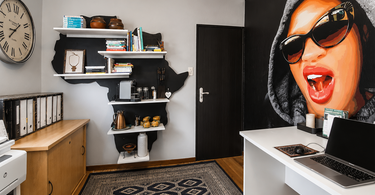 Wide view of a home office with a large custom portrait mural on a black wall, styled with a white desk, bookshelves, wood flooring, and patterned rug.