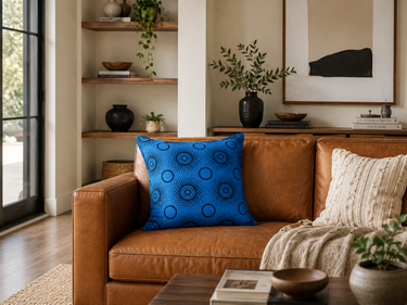 Sky-blue NIZARRA Setswana-inspired cushion with circular dark blue motifs styled on a warm brown leather sofa in a modern living room.
