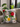 Grey Tsonga cushion with red, yellow, pink and blue flowers on grey and white striped fabric, styled on a neutral sofa near indoor plants and a window.