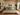 Green Sepedi-inspired NIZARRA cushion styled at the centre of a modern neutral bedroom with layered green bedding and indoor plants.