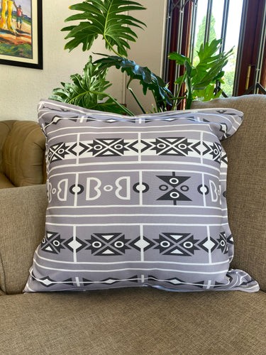 Grey cushion with a black and white Bapedi-inspired geometric pattern styled on a lounge chair beside leafy plants.