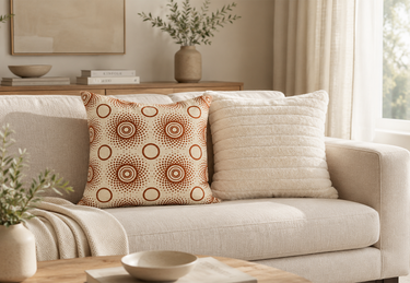 Cream and rust-brown Setswana-inspired patterned cushion styled on a neutral sofa with soft textured cushions in a calm modern living room setting.