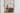 A minimalist Afro-modern interior viewed through white double doors, featuring a Black man working on a laptop at a stone table under a soft pendant light, set against a refined NIZARRA-style geometric wallpaper in warm neutral tones with subtle cultural patterning.