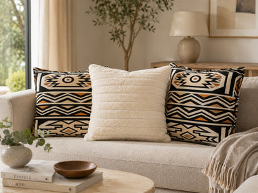Brown Ndebele-inspired NIZARRA cushions styled with a cream textured cushion on a neutral sofa in a warm Afro-modern living room.