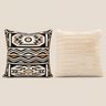 Two square throw pillows on a solid beige background. The left pillow features a Ndebele geometric pattern in black, cream, and ochre orange.Ndebele-print cushion cover in black, cream and ochre displayed beside a cream ribbed faux-fur cushion cover.