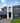 Wide view of a modern residential façade featuring grey and white walls, black doors, custom entrance shade, striped window frosting, and a clean front lawn.