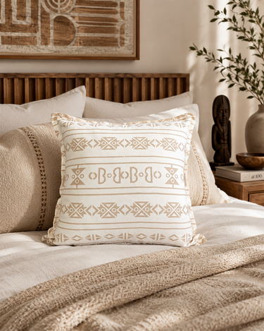 Cream Sepedi-inspired NIZARRA cushion styled on a layered neutral bed with warm wood details and textured bedding.