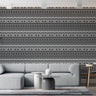 Swazi inspired wallpaper grey background with geometric repeat pattern of zig zag lines and shield and spear graphics and oval shapes in colours black and white. Setting is a living room with a corner sofa that complement the wallpaper.