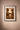 Framed African artwork with a wide off-white mount and black frame on a textured plaster wall, depicting two figures exchanging a vessel in warm ochre tones.